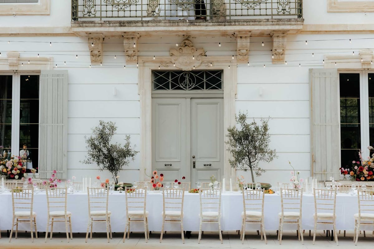 Long banquet tables with white linens and coral floral centerpieces set before a French chateau with sage-green shutters