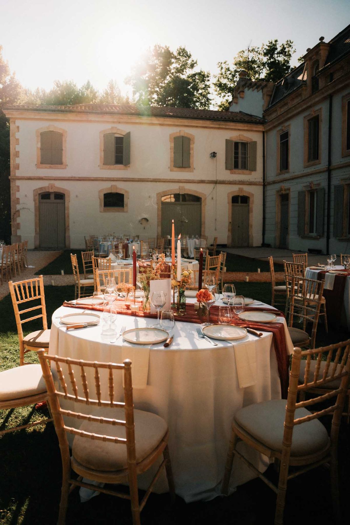 Round terracotta-runner table with orange roses, burgundy tapers, and gold chiavari chairs at golden hour