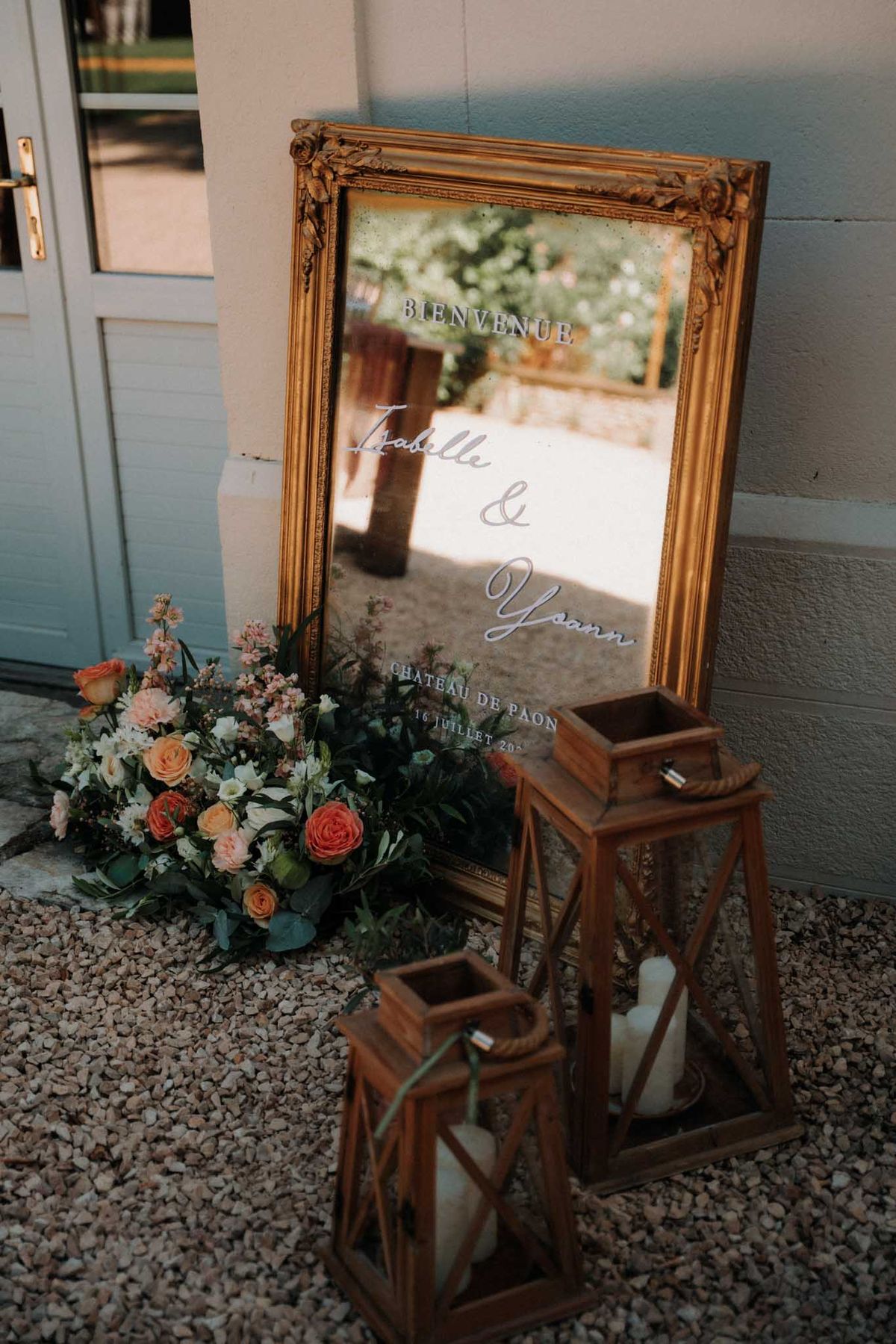 Gold-framed Bienvenue mirror sign with coral roses, wooden lanterns, and pillar candles on gravel