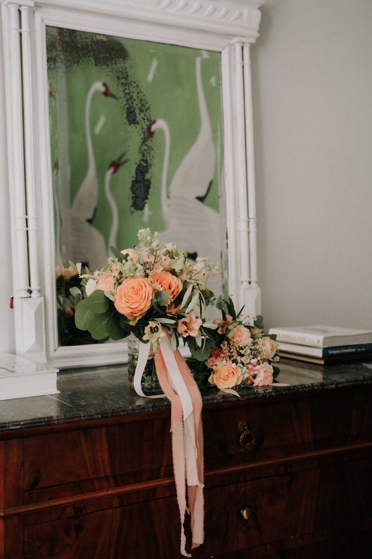 Peach rose and blush spray rose bouquet with trailing silk ribbons on marble-topped dresser beside foxed mirror