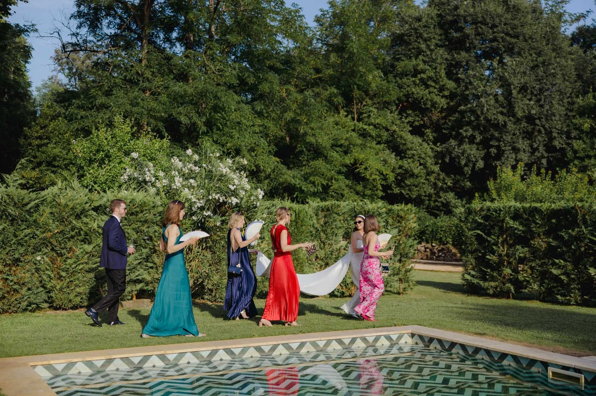 Six guests in colourful gowns walking across lawn beside chevron-tiled pool with hand fans at garden cocktail hour
