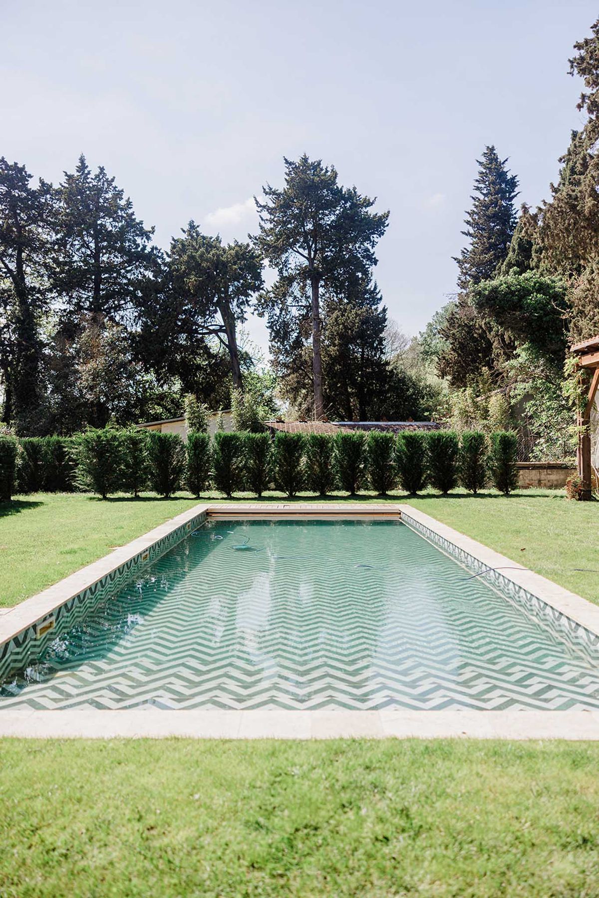 Rectangular swimming pool with green and white chevron tile pattern trimmed topiary hedge and wooden pergola