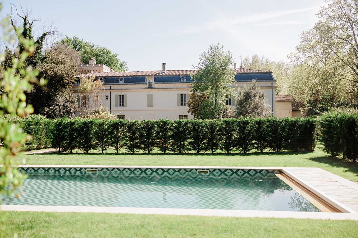 Cream manor with mansard roof and green shutters behind chevron-tiled pool and clipped formal hedge