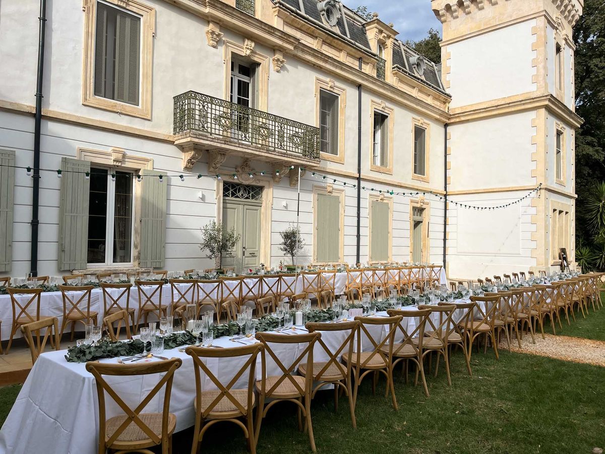 Long tables with greenery runners, olive tree centerpieces, and coloured string lights along chateau limestone facade