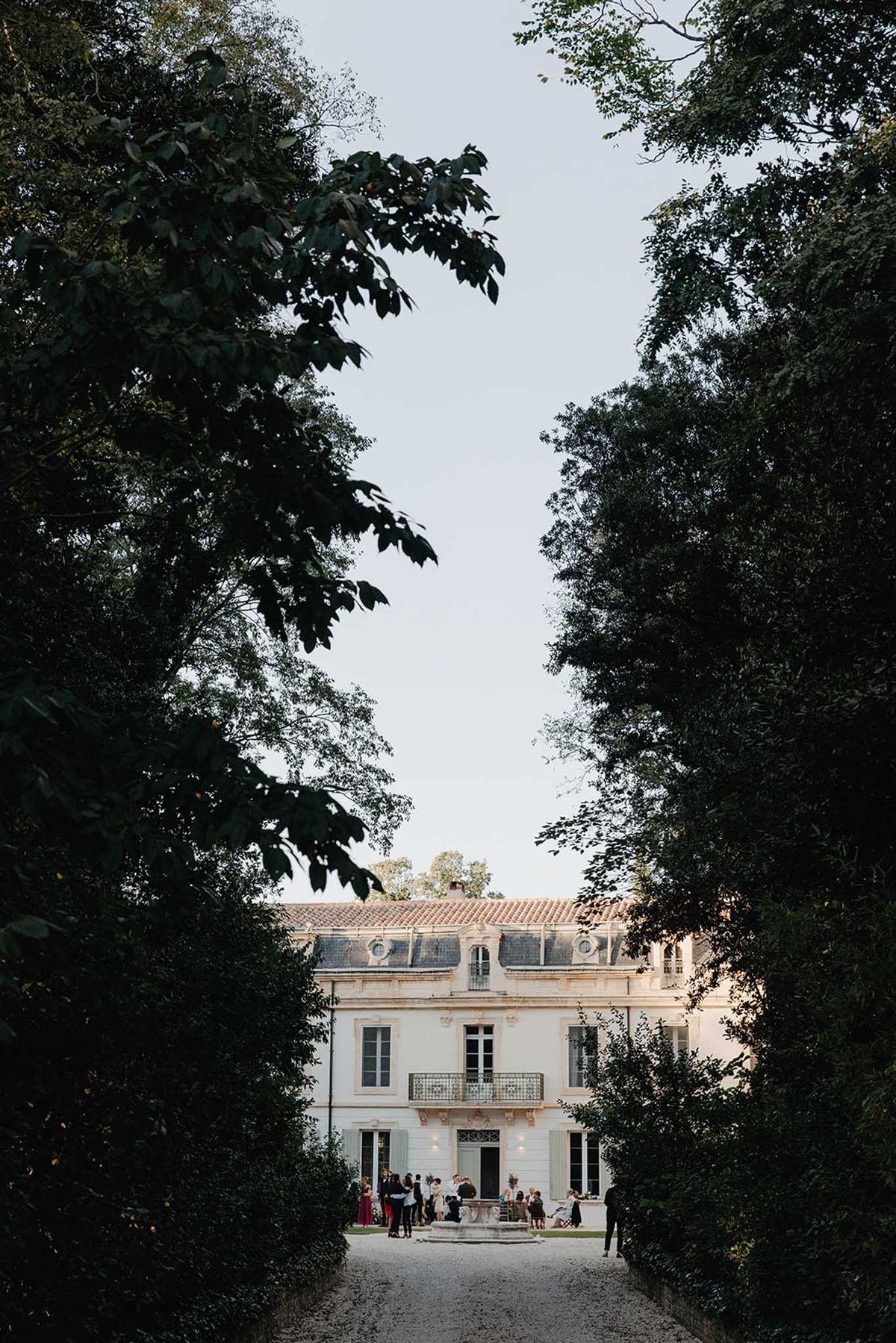Tree-lined gravel driveway at dusk with guests at fountain before white stone chateau with blue shutters