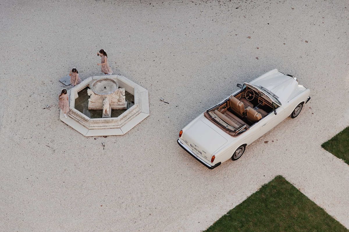 Aerial view of three flower girls in blush tulle dresses gathered around octagonal fountain beside vintage cream convertible