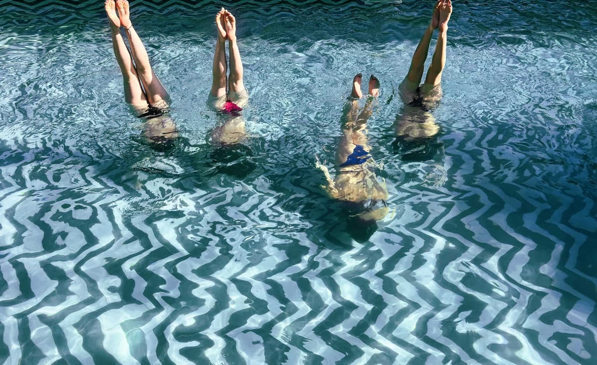 Five people submerged in pool with bare legs visible above turquoise water in playful synchronized pose