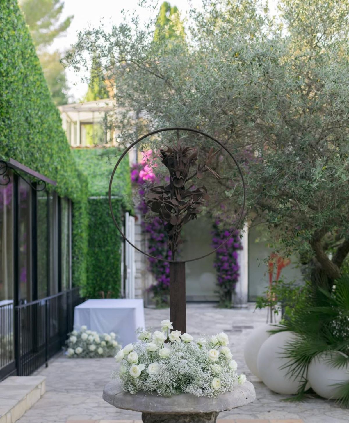 Rust metal botanical sculpture on pedestal with white roses and gypsophila beside bougainvillea wall