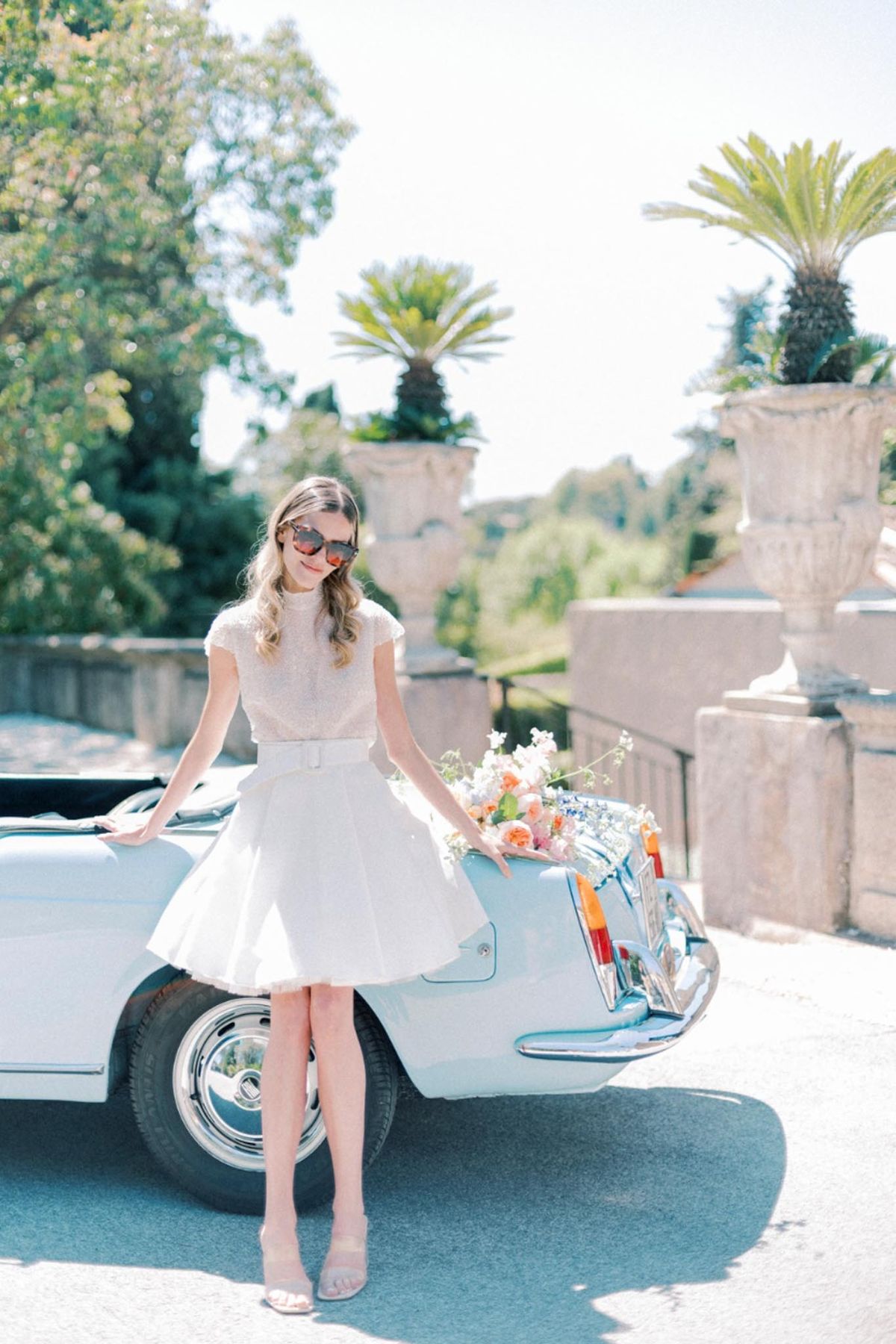 Bride in embellished crop top and midi skirt leaning against vintage white convertible with peach rose bouquet at chateau