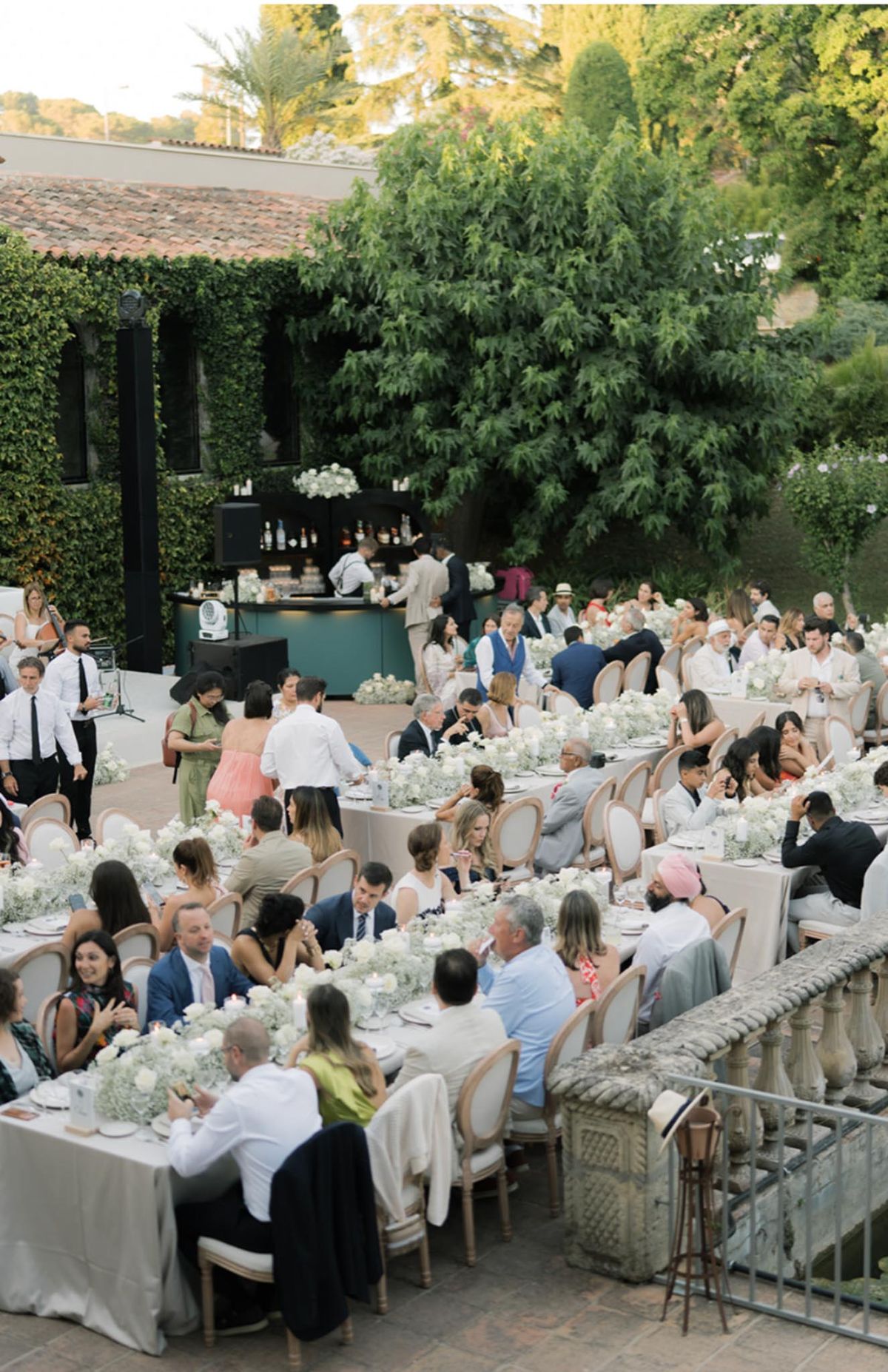 Sixty guests at courtyard reception with all-white babys breath centerpieces Louis XVI chairs teal bar and live guitarist