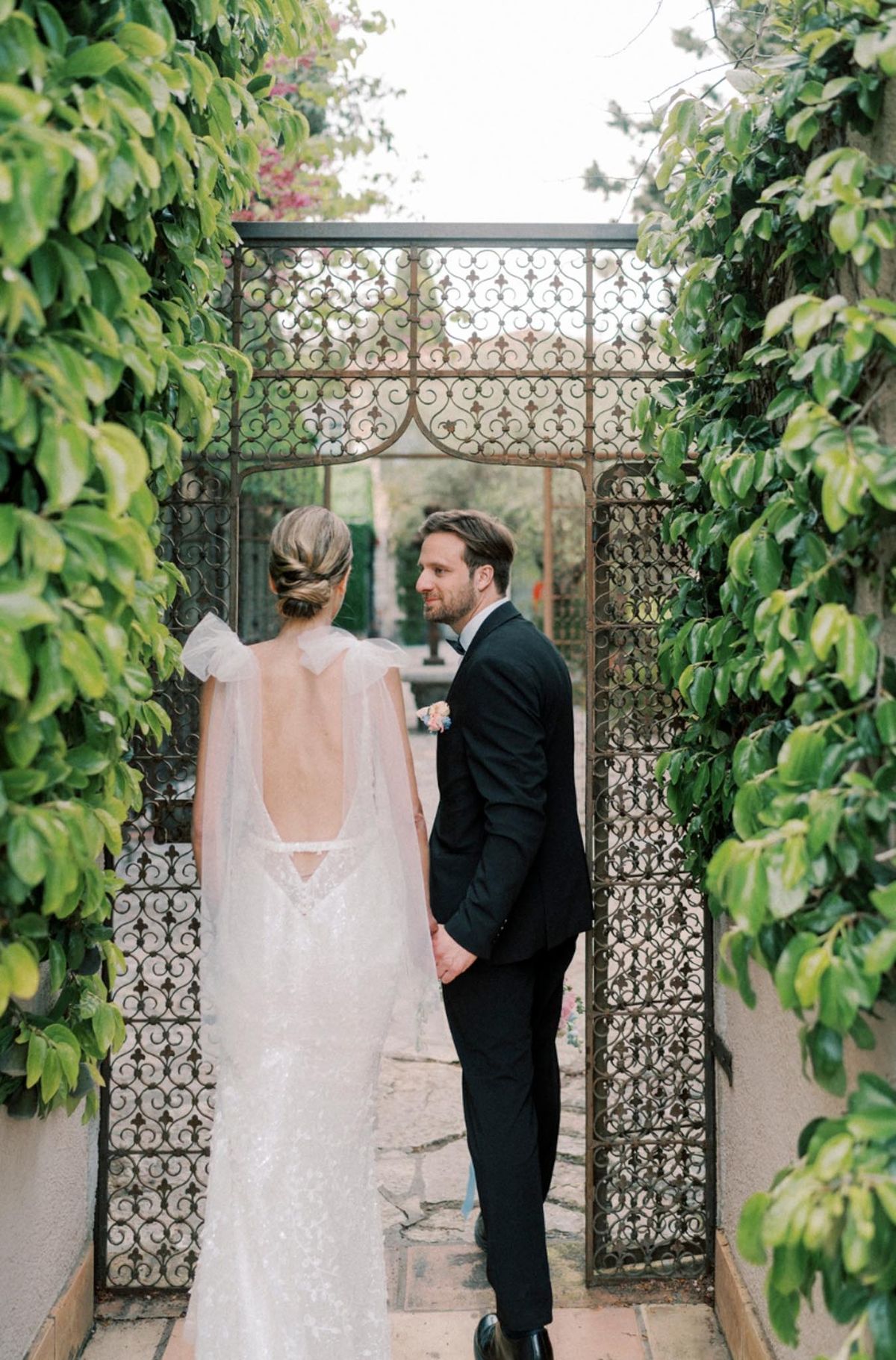Couple framed by wrought-iron gate showing bride's sequined gown open back and tulle cape