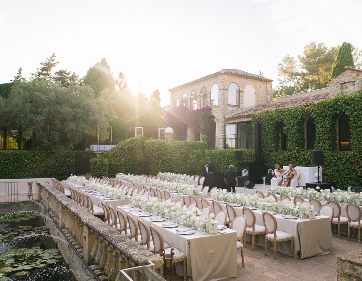 Outdoor reception on stone terrace with long banquet tables, white floral runners, and string ensemble at golden hour