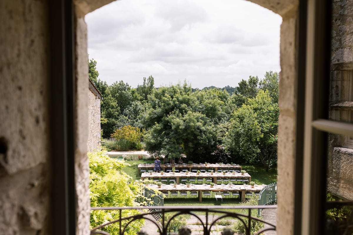 U-shaped farm tables with potted plant centerpieces and sage chairs viewed from chateau window