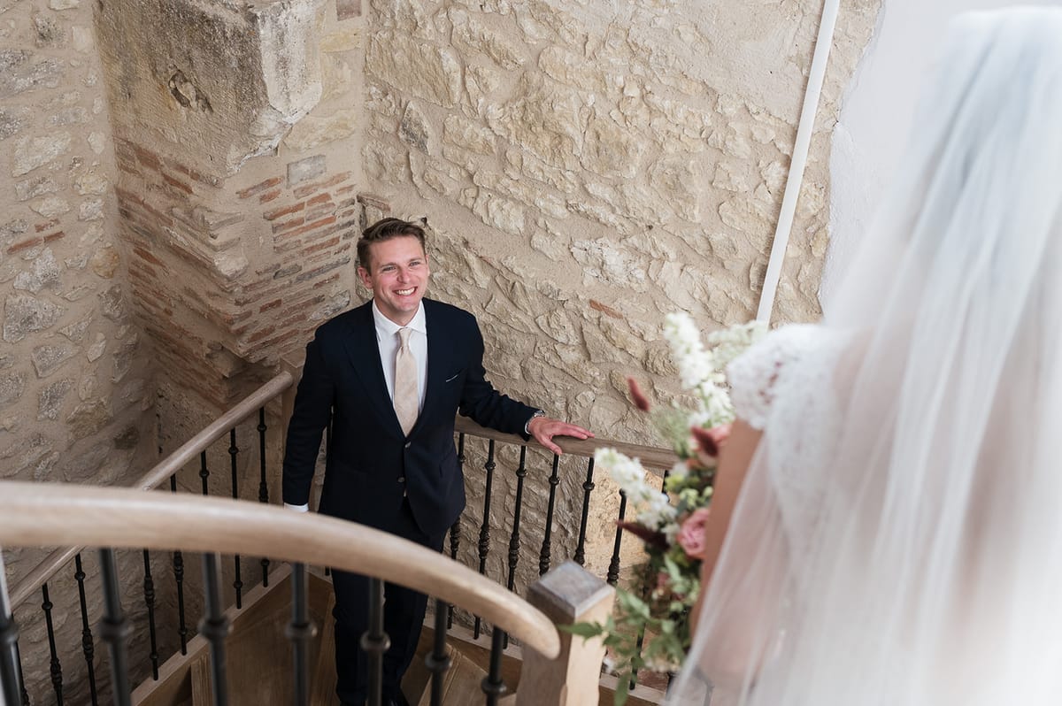 Groom looks up smiling at bride descending stone staircase with bouquet in soft focus from above