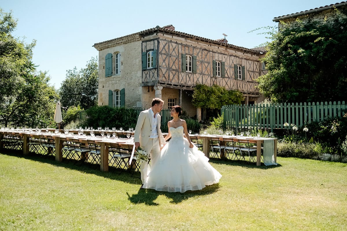Bride in ivory ballgown and groom in cream linen suit walking across lawn in front of timber-framed manor house