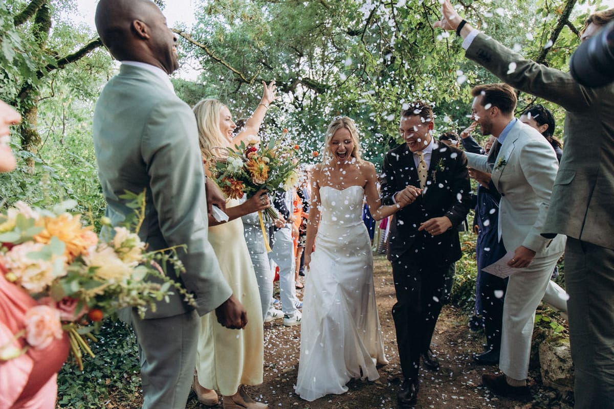 Bride and groom walking through white petal confetti tunnel with guests, bridesmaids carrying orange and rust bouquets