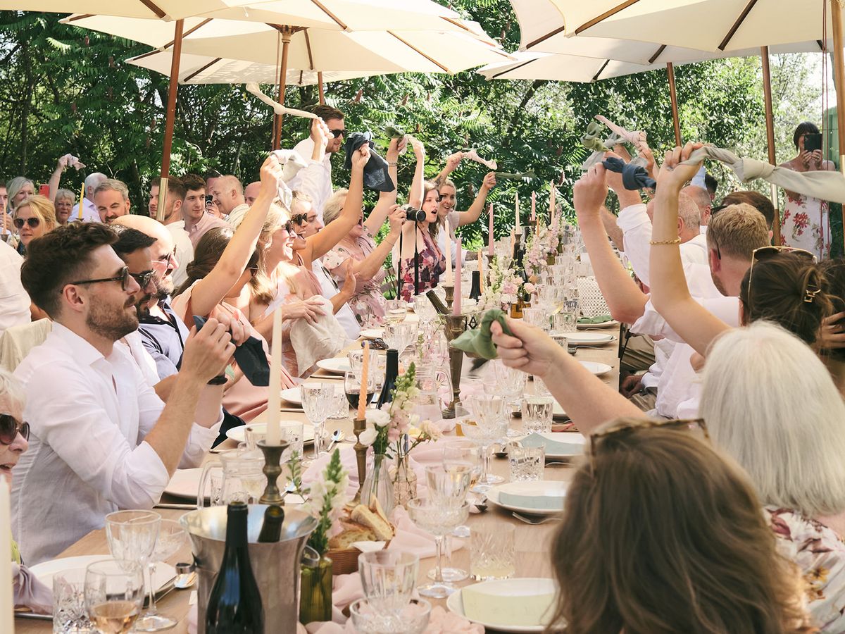 Guests waving sage napkins at long garden table with blush runner, pink taper candles, and cream parasols
