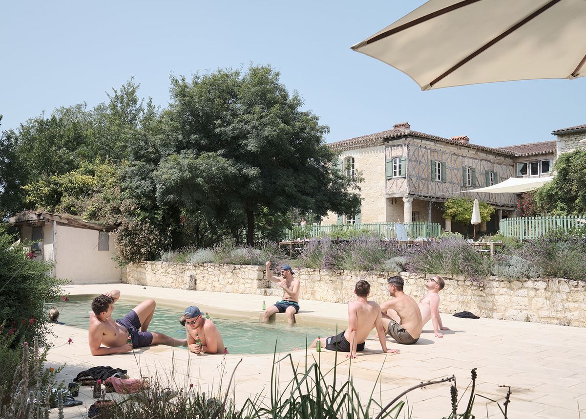 Guests relaxing by small pool with lavender border at half-timbered French manor with green shutters in summer sun