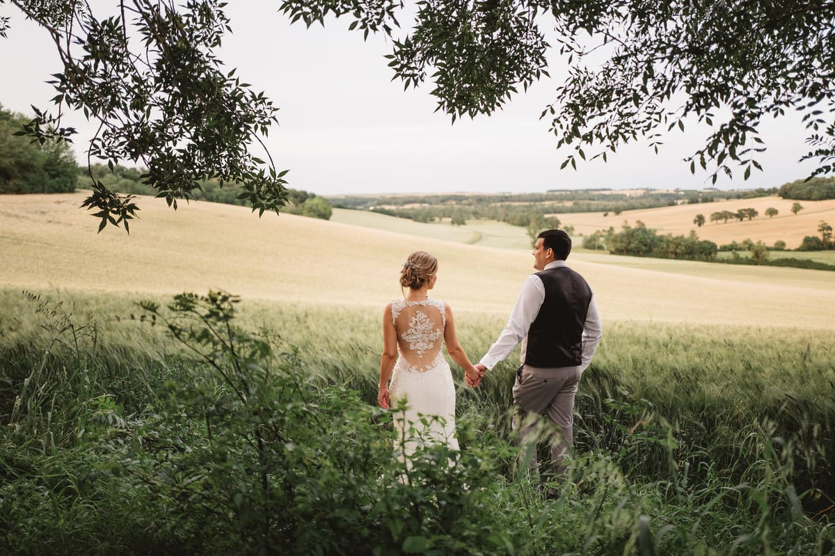 Bride in ivory lace-back gown and groom in navy waistcoat walking hand in hand through wild grass in the French countryside