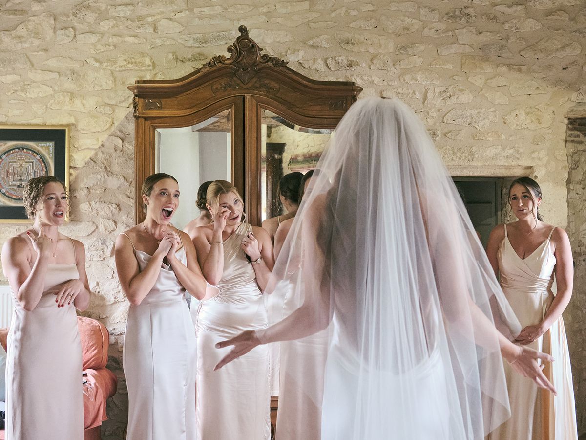 Bridesmaids in blush satin dresses reacting with tears and applause as bride reveals her gown