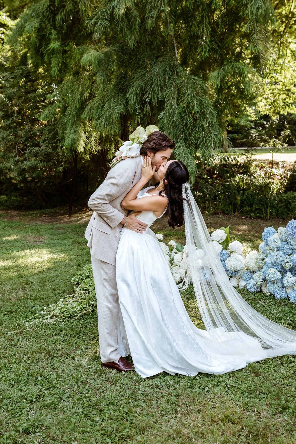 Groom dipping bride for a kiss in garden setting with cathedral veil, white orchid bouquet, and blue hydrangea arrangements