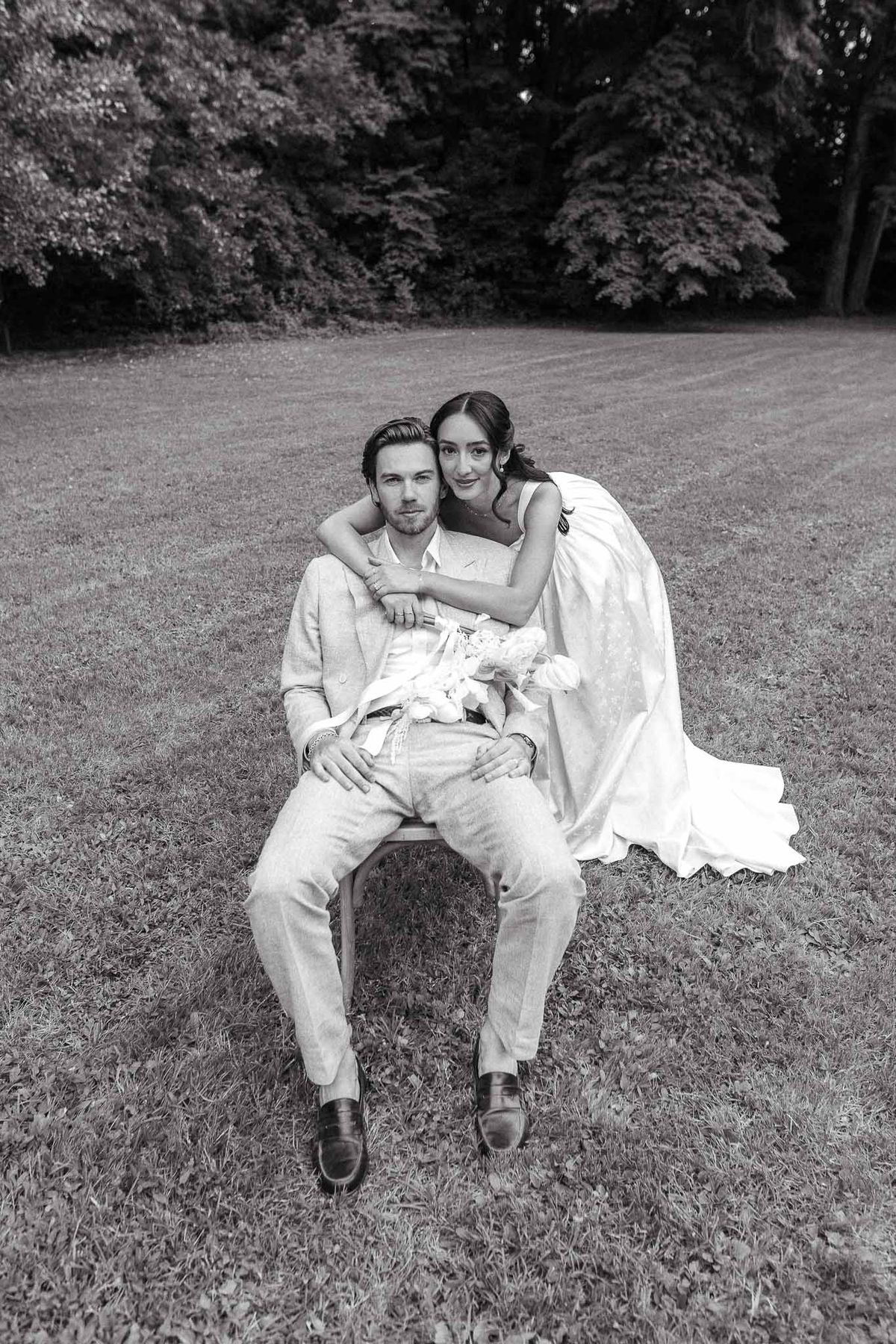 Black and white portrait of groom seated with bride standing behind, arms draped, on manicured lawn