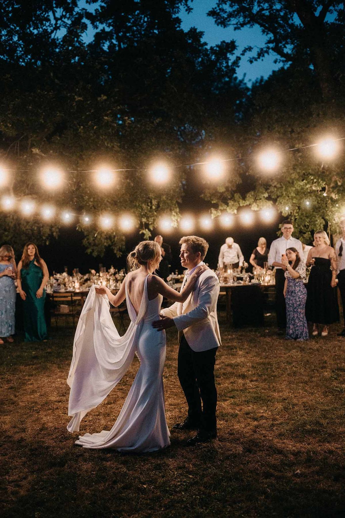 First dance at dusk on grass lawn with festoon lights bride in white cape gown groom in ivory blazer
