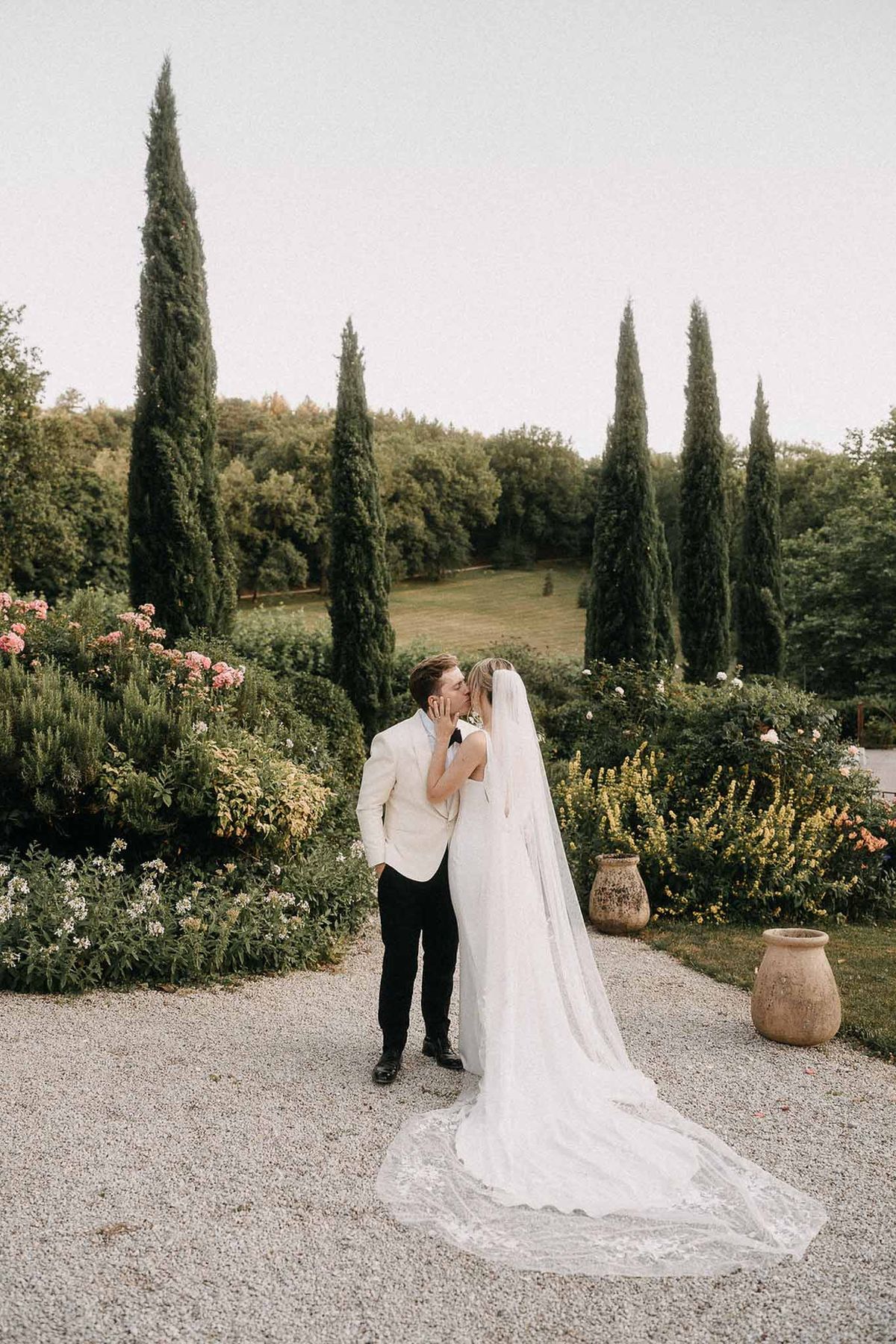 Couple kissing in formal garden with lace-edged veil fanning on gravel, pink roses, and cypress trees