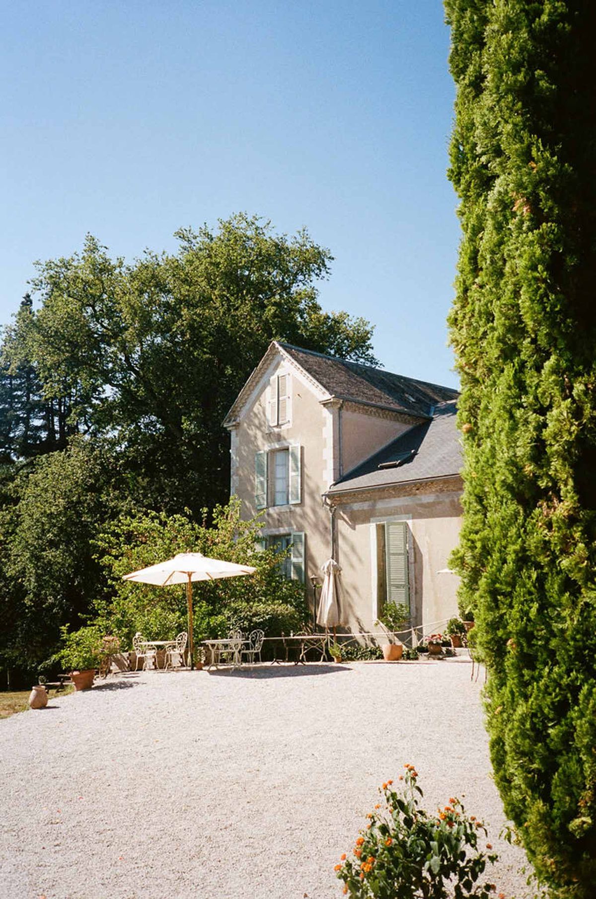 Cream manor with pale blue shutters, wrought-iron bistro seating, parasol, and cypress on gravel courtyard