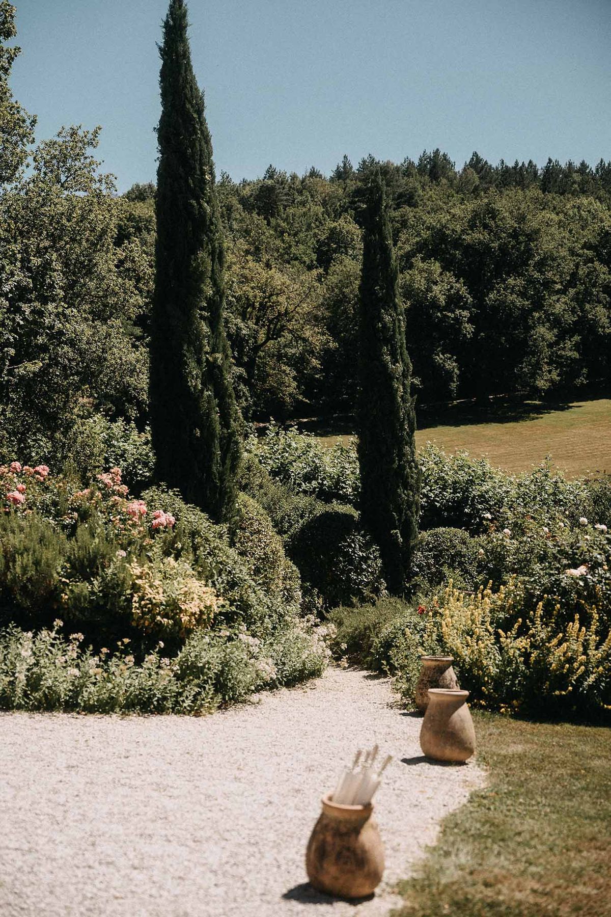 Gravel pathway lined with cypress trees, pink roses, and terracotta urns in Provencal venue garden