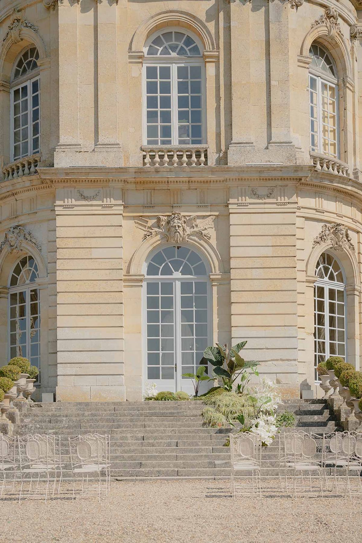 Pre-ceremony setup with white wrought-iron chairs facing golden limestone chateau steps with white lily arrangements
