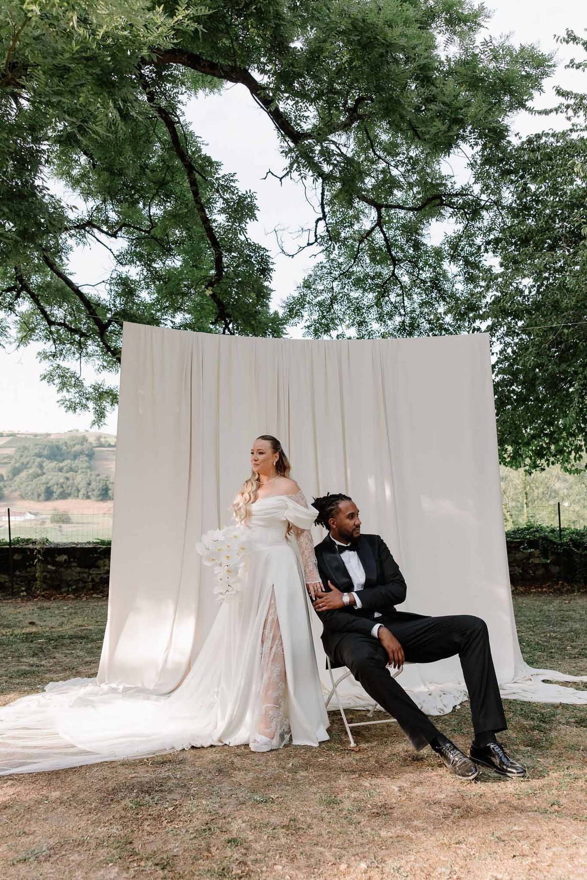 Editorial couple portrait against ivory drape outdoors bride in puff-sleeve gown with orchid bouquet groom seated in tuxedo