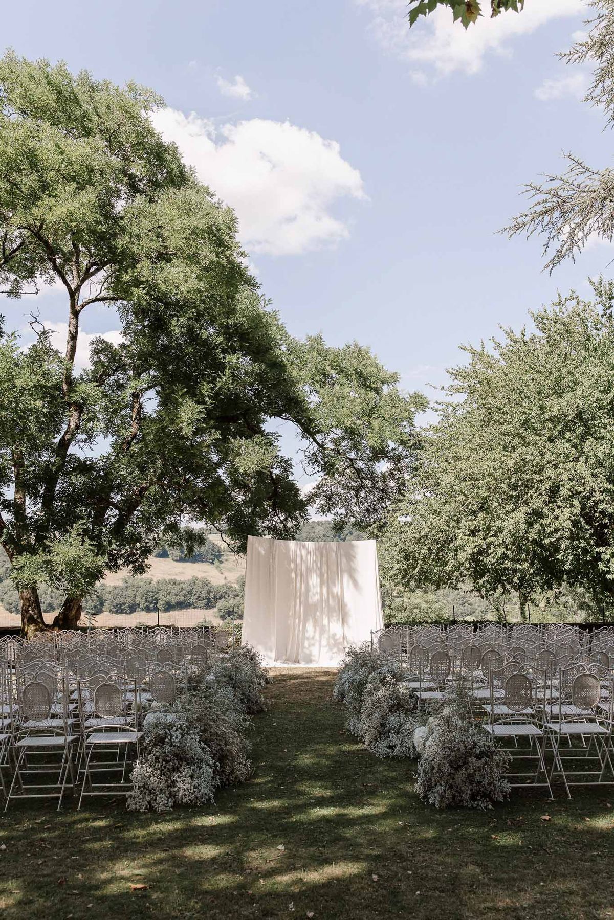 Ceremony setup with white draped altar, baby's breath aisle borders, and white metal chairs on lawn