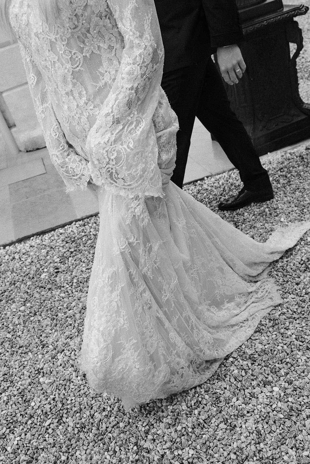 Black and white close-up of lace wedding gown train trailing across gravel beside groom shoes