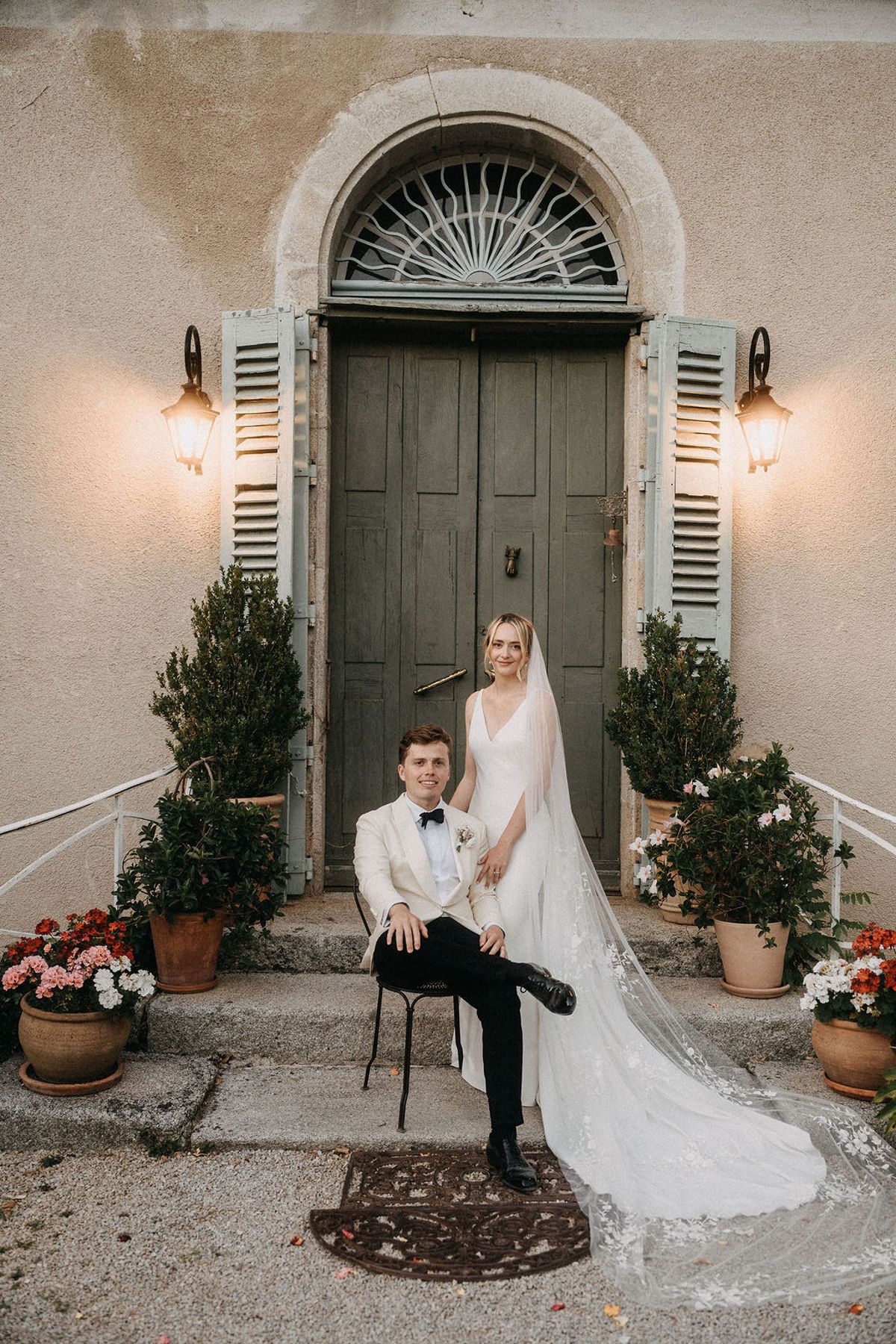 Bride in sleeveless white gown with cathedral lace veil standing behind seated groom at chateau entrance with green door