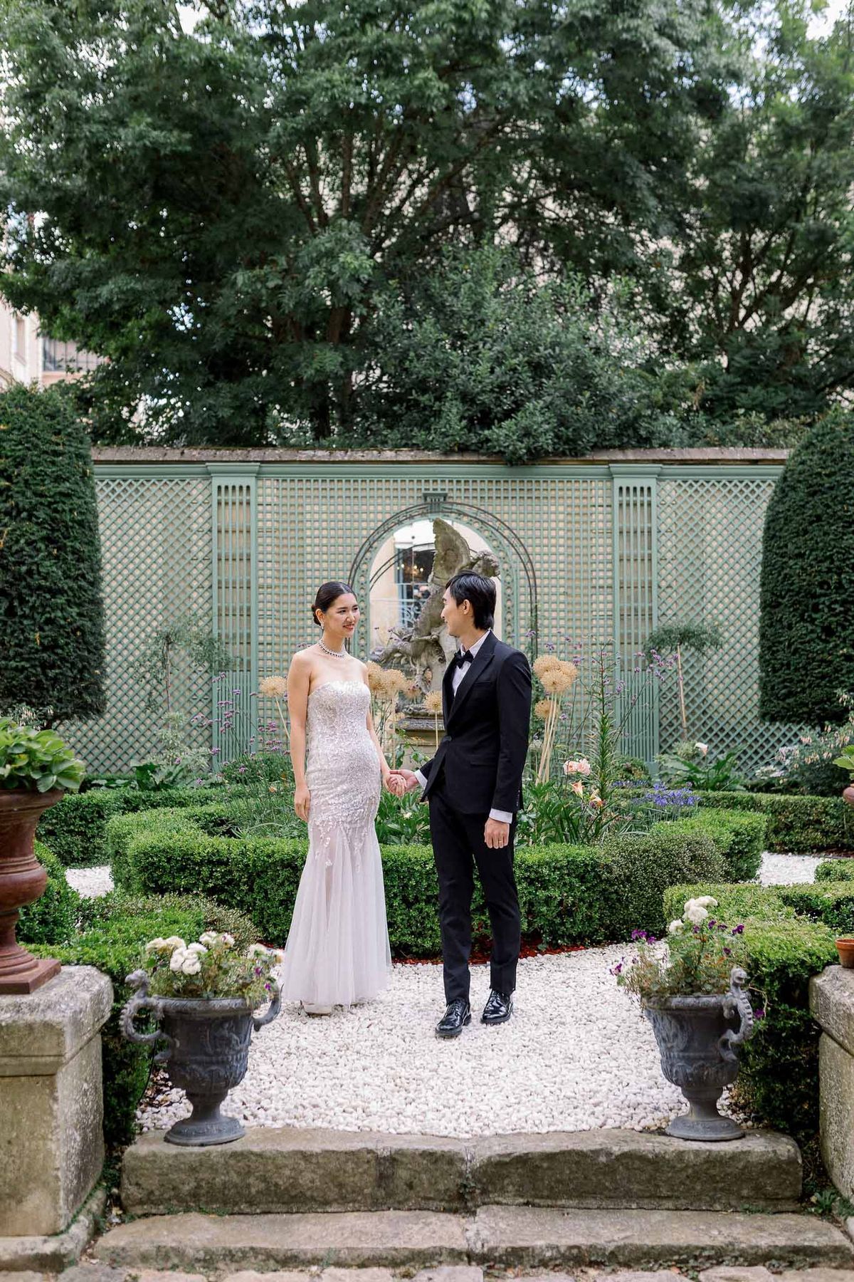 Couple holds hands on gravel path in formal garden with boxwood parterres and sage lattice trellis behind