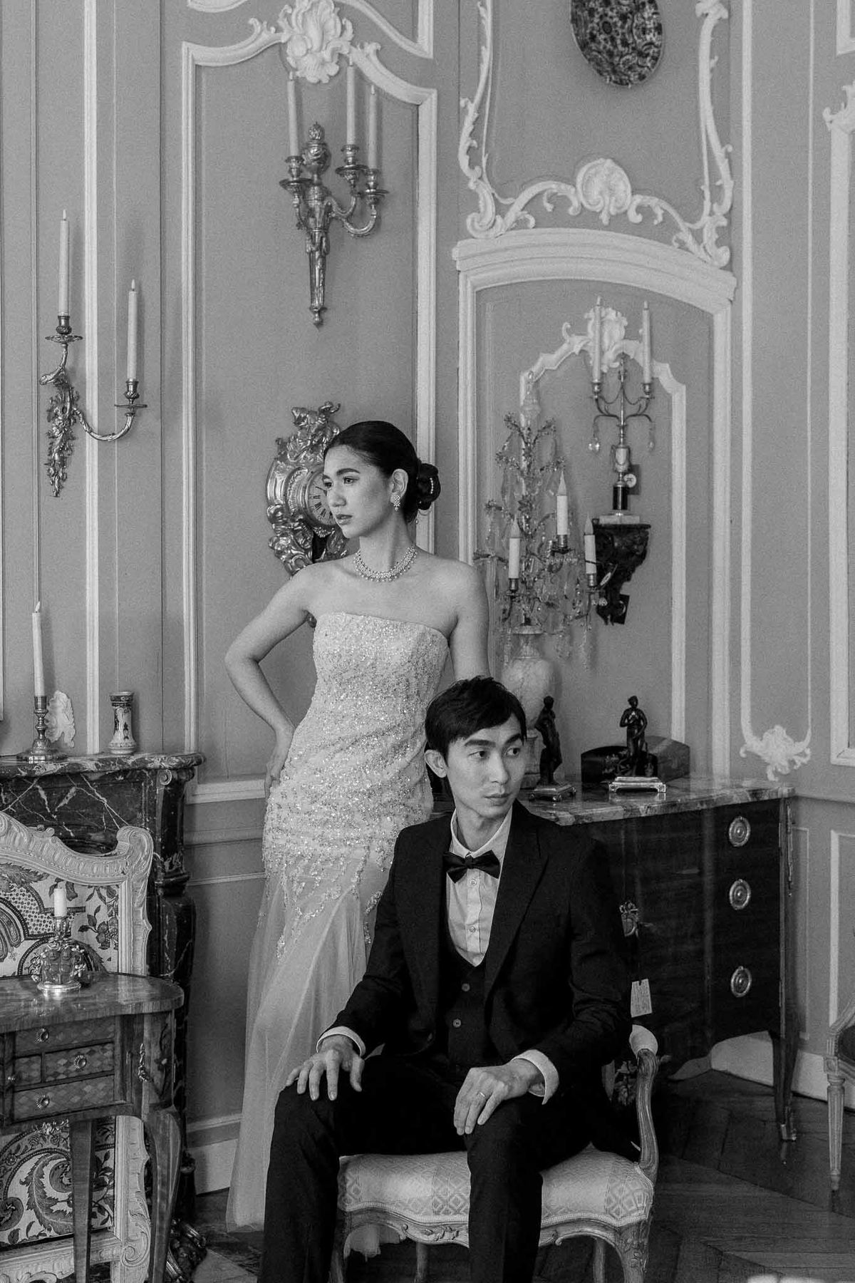 Black and white bride standing behind seated groom in Rococo salon with gilded sconces and ornate plasterwork
