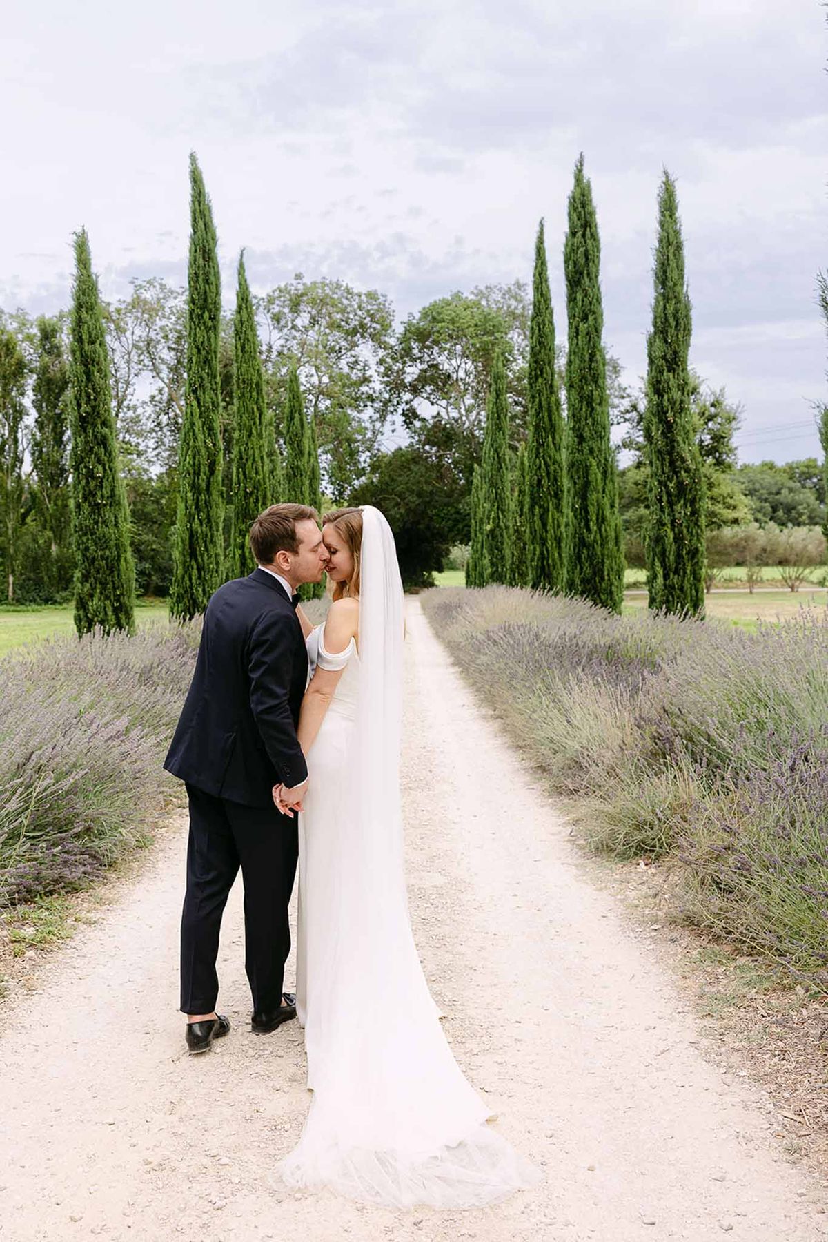 Bride and groom kissing on lavender-lined gravel path with cathedral veil trailing behind and cypress trees beyond