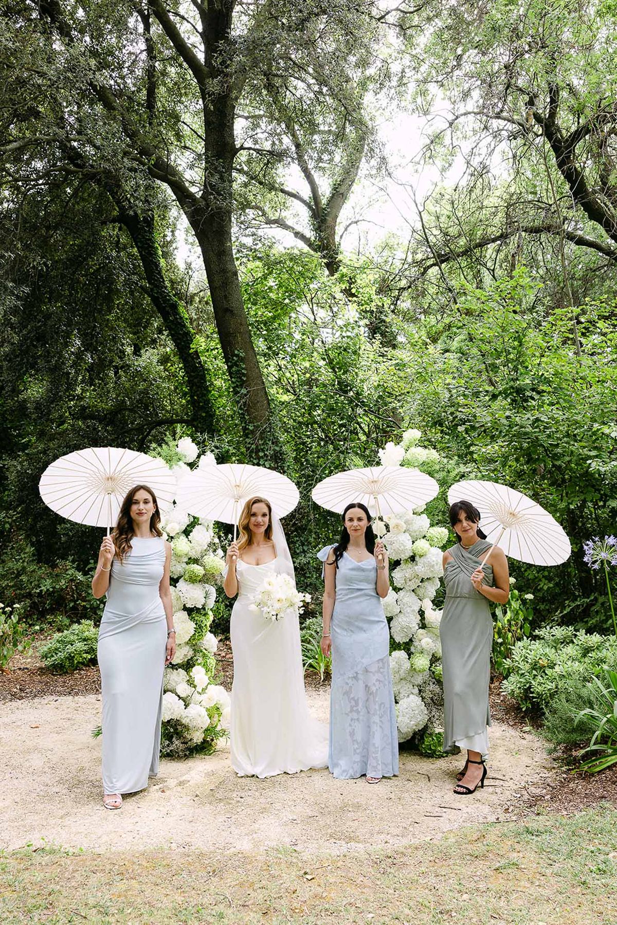 Bride and three bridesmaids holding white parasols beside a white hydrangea floral installation