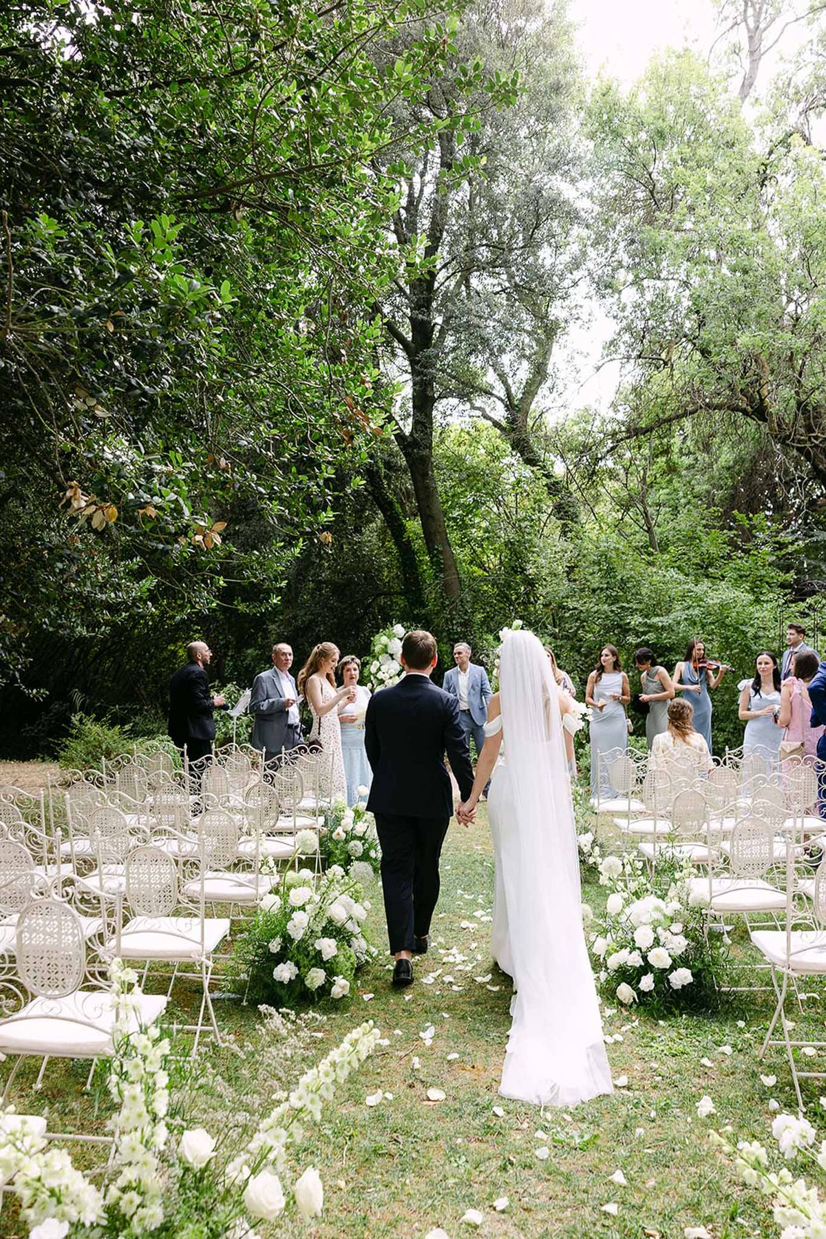 Couple walks back down petal-strewn grass aisle past white rose arrangements with bridesmaids in dusty blue