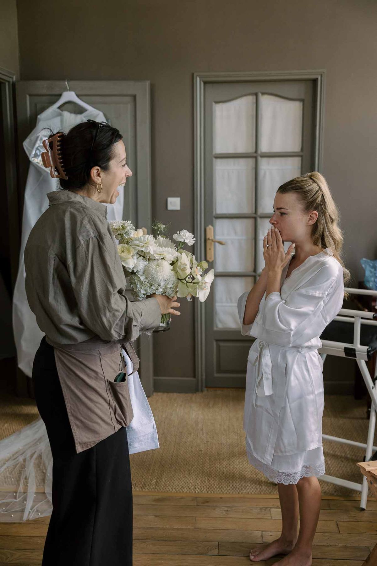 Bride in white robe reacting emotionally as florist presents white and green bridal bouquet