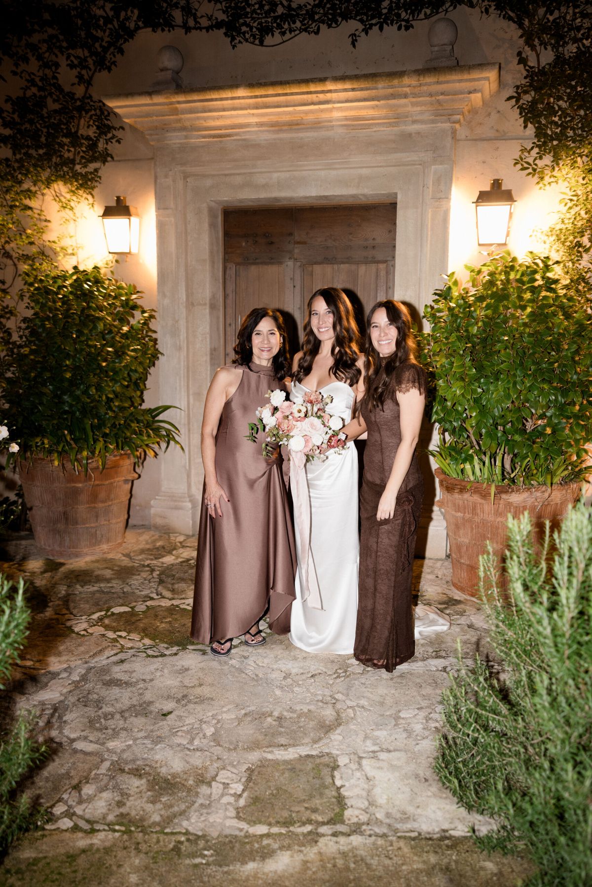 Bride in white satin gown with two companions in mauve and brown dresses at cobblestone entrance