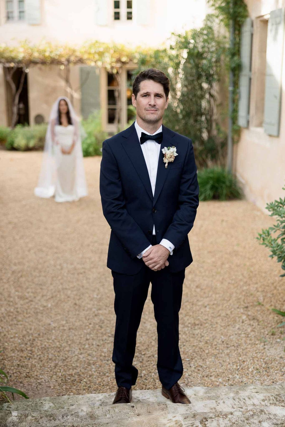 First look with groom in navy tuxedo waiting as bride approaches in soft focus at Provencal courtyard