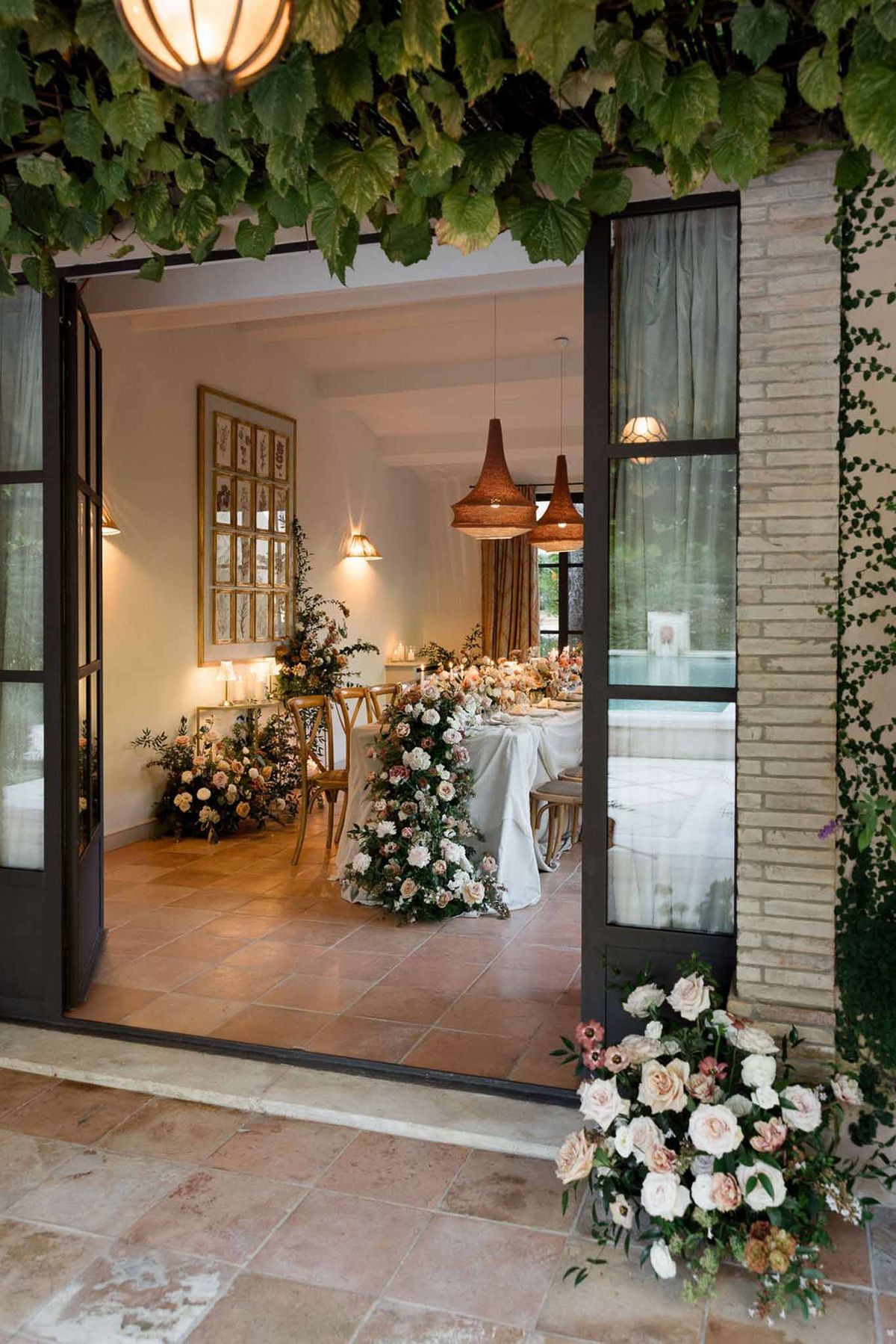 Indoor reception through open doors with blush and apricot rose runner, rattan pendants, and cascading floral installation