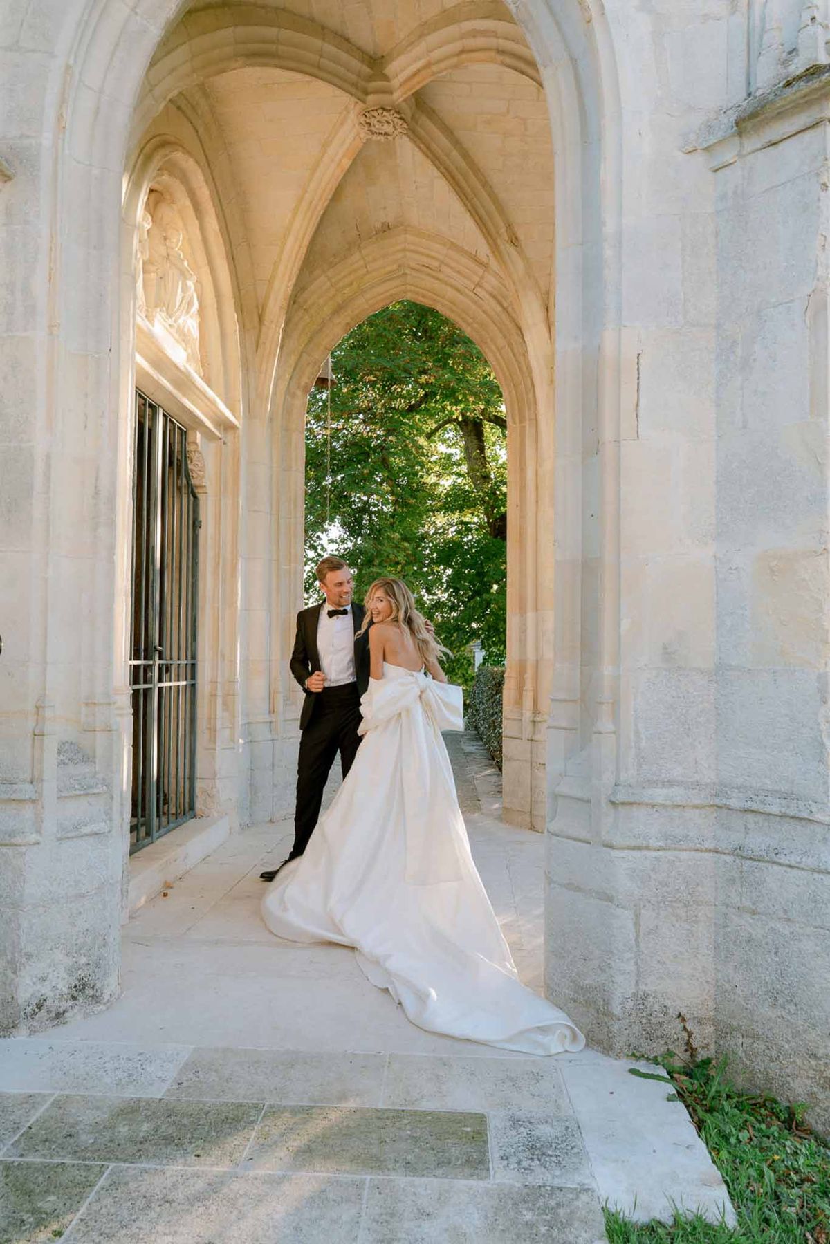 Bride in strapless ballgown with bow detail and groom in black tuxedo under Gothic stone archway at golden hour