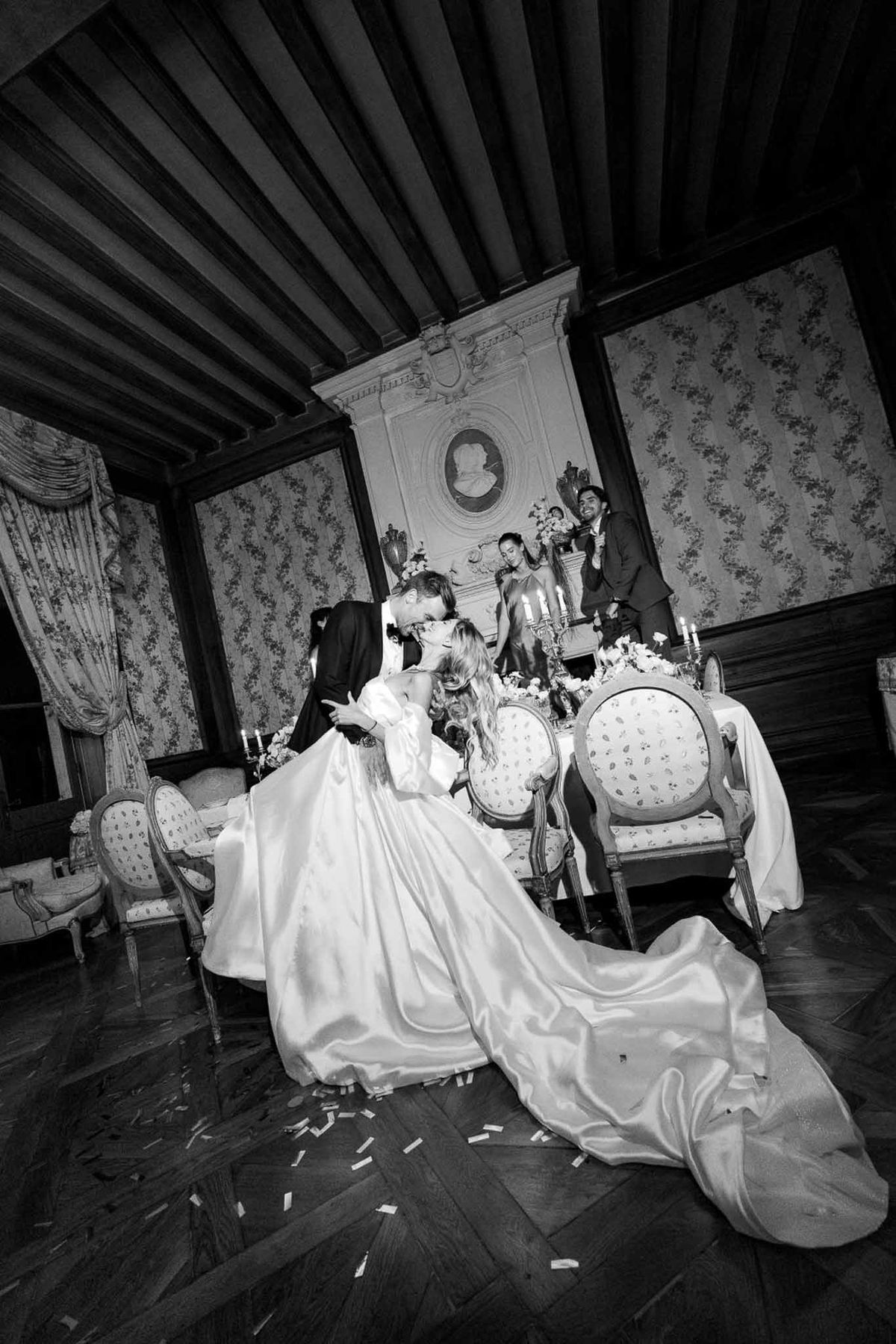 Couple kisses at sweetheart table with confetti-strewn parquet in coffered-ceiling chateau salon in B&W