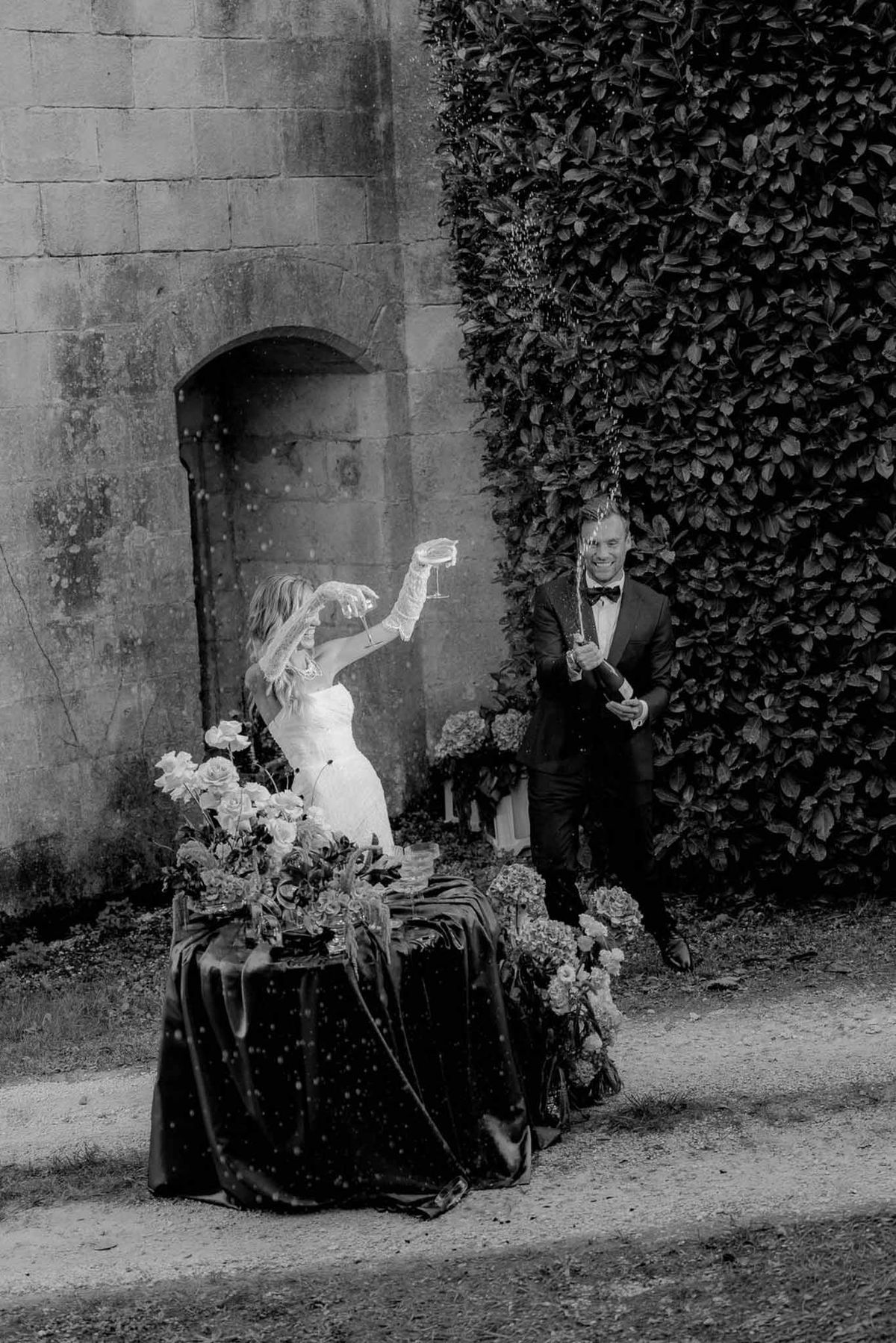 Black-and-white image of bride and groom spraying champagne bottle outside chateau with rose and hydrangea arrangement