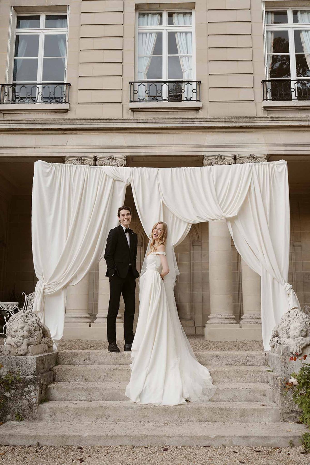Laughing couple on chateau steps with ivory draped arch, bride in cathedral veil and off-shoulder gown