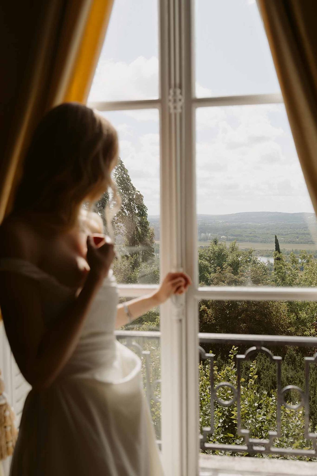 Bride in white dress looking out tall French window with gold curtains over valley landscape
