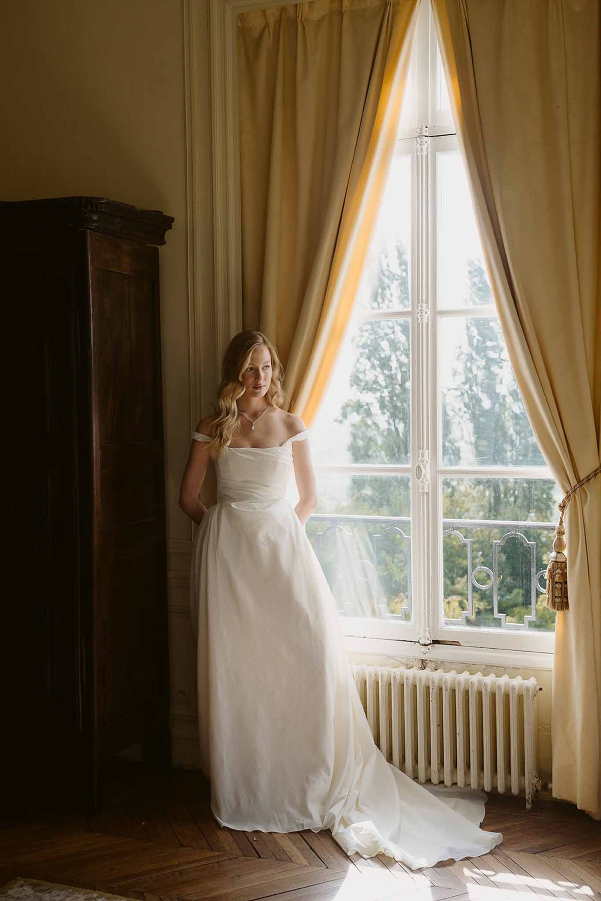 Bride in off-shoulder ivory gown beside tall window with gold curtains in chateau room with herringbone floor
